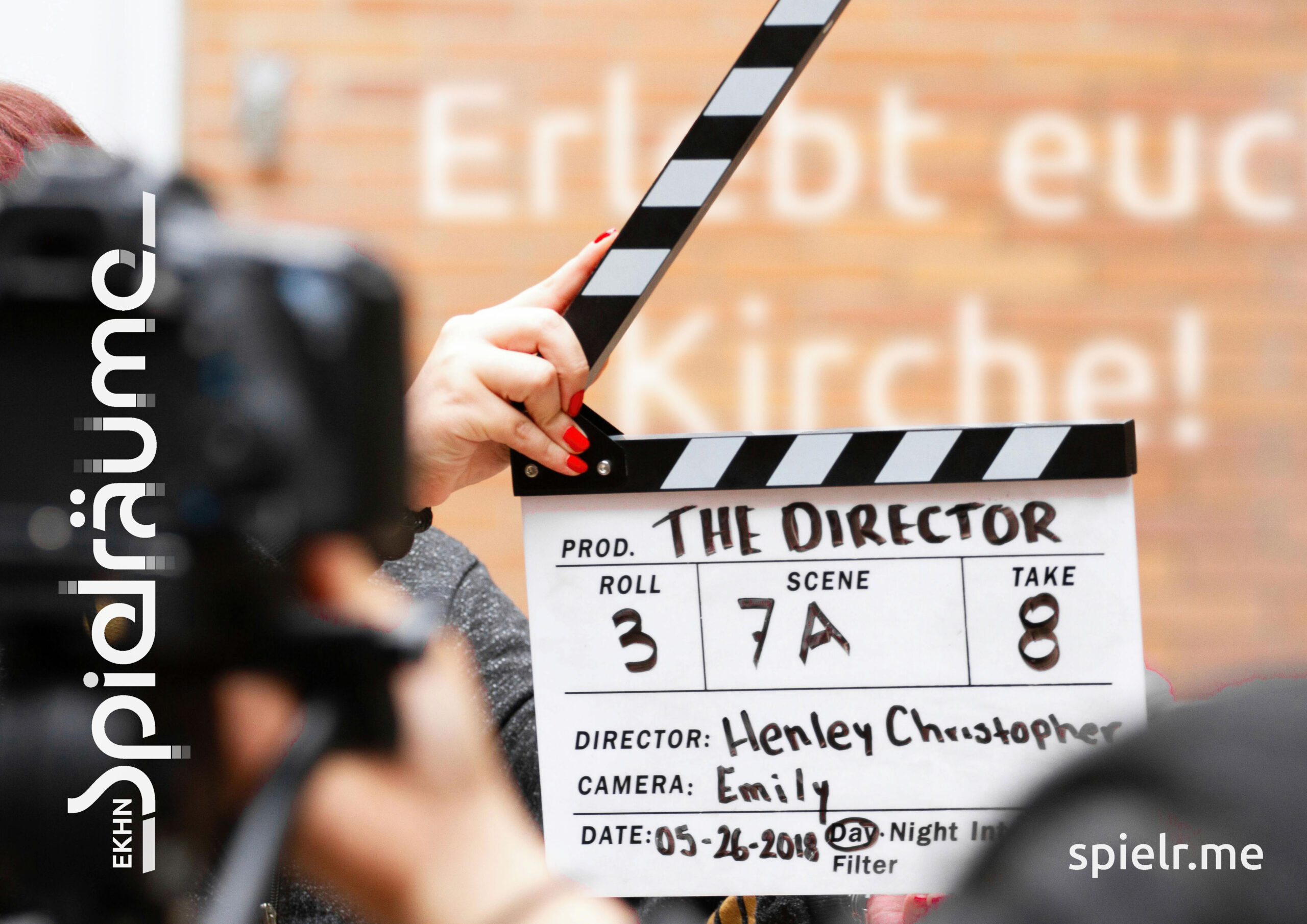 Eine Filmklappe mit der Aufschrift THE DIRECTOR wird vor eine Kamera gehalten; im Hintergrund sind unscharfe Hände und eine Backsteinmauer zu sehen, über die sich ein Text legt.