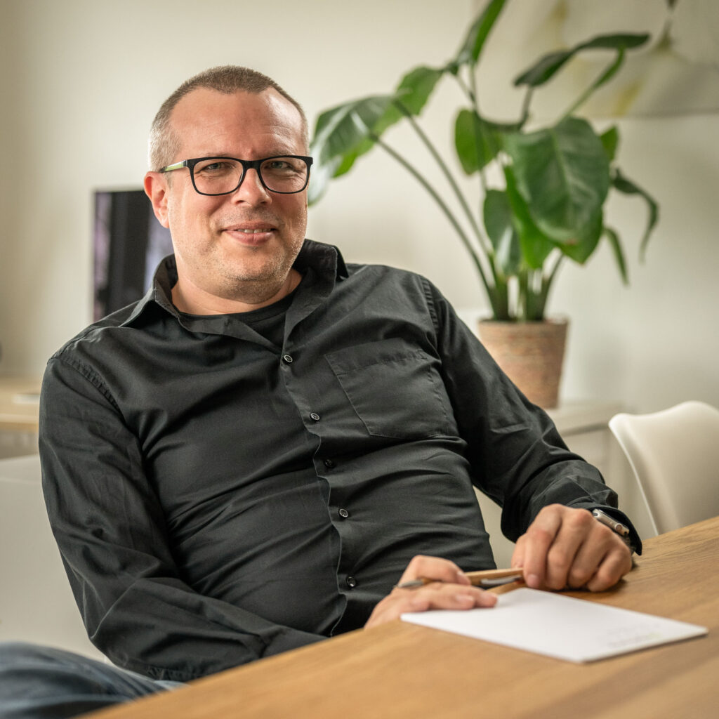Ein Mann in einem schwarzen Hemd mit Knöpfen und einer Brille sitzt mit Stift und Papier an einem Holztisch.